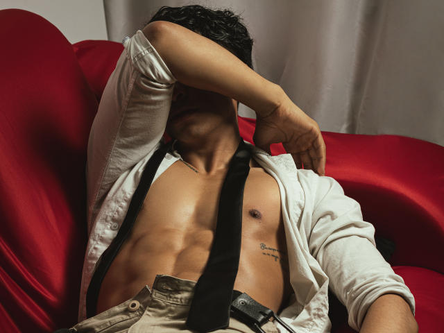 ApoloCrus, sem camisa, com a camisa branca aberta, gravata preta e calças bege, está sentado em uma cadeira vermelha, com a mão cobrindo o rosto, revelando abdominais definidos e uma pequena tatuagem de um mamilo..