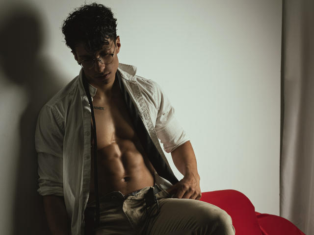 ApoloCrus, con cabello rizado, usando gafas, camisa blanca abierta, mostrando un cuerpo tonificado, musculoso y bronceado con abdominales visibles, sentado en una silla roja, con los pantalones ligeramente bajados, mirando hacia abajo..