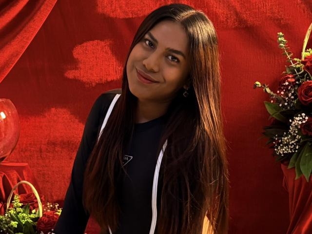 KimmySolange with long brown hair, black top, white straps, smiling at camera, red background, bouquet of flowers and glass vase on left, soft light, cute, attractive, sexy.