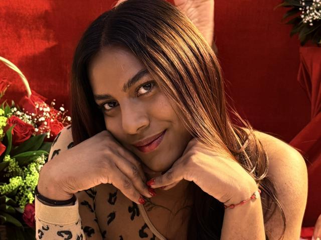 KimmySolange with long brown hair, red lipstick, and a leopard print top, smiling with hands under her chin, red nails, and a red bracelet, in a sunny, vibrant setting.