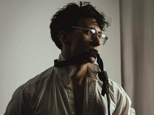 ApoloCrus, dark curly hair, glasses, white button-down shirt, black necktie hanging from mouth, side profile, serious expression, dim lighting, medium shot, subtle shadows.