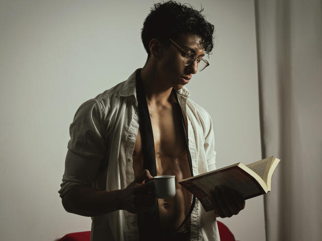 ApoloCrus, torse nu, avec des cheveux noirs bouclés et des lunettes, tenant un livre et une tasse blanche, portant une chemise blanche ouverte, révélant une poitrine musclée, tonique et séduisante..