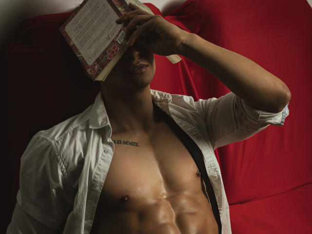 ApoloCrus, avec un livre sur le visage, chemise ouverte, dévoilant des abdominaux sculptés et un tatouage sur la poitrine, allongé sur un canapé rouge, paraissant sexy et détendu..