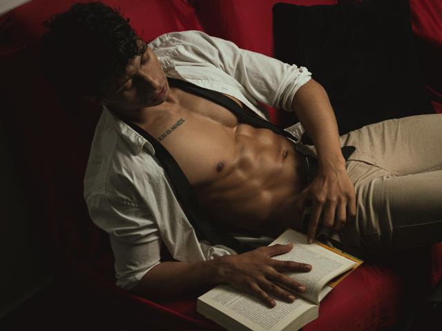 ApoloCrus, senza camicia, muscoloso, con un tatuaggio sul petto, sdraiato su un divano rosso, che legge un libro, indossa pantaloni beige, una camicia bianca aperta, una mano sull'inguine, capelli scuri..