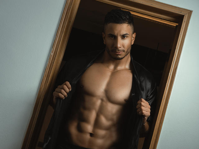 ZaneMaverick, senza camicia, con un torso muscoloso e scolpito, giacca di pelle nera aperta, in piedi in una porta, capelli corti e scuri, espressione seria, pelle media, addominali visibili, che guarda la fotocamera..