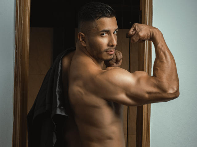 ZaneMaverick, un homme musclé, torse nu, à la peau foncée, avec une mâchoire forte et les cheveux courts, contracte son biceps droit, exhibant son bras et sa poitrine musclés et bien définis..