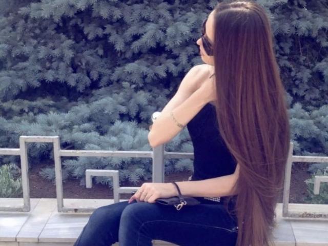 MalaniSweet with long brown hair, black sleeveless top, and black jeans, sitting on a wooden bench, wearing sunglasses, arm bracelet, and black wristband, against a tree-filled background.