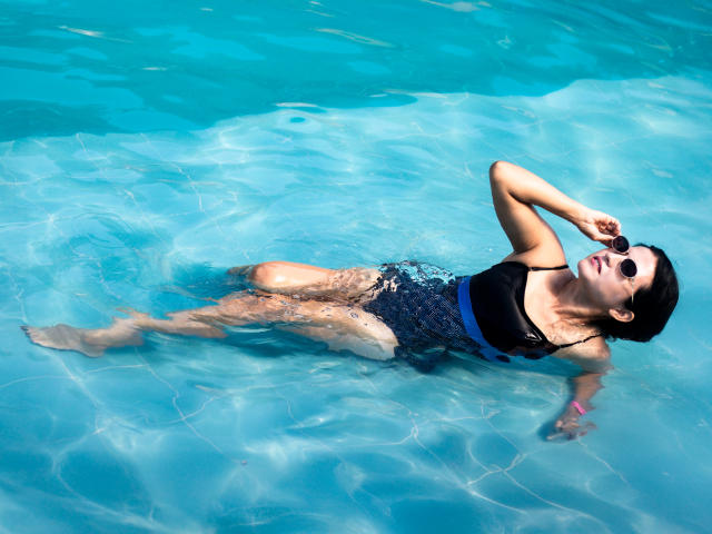 RenataRivas floating in a pool, wearing a black one-piece, blue patterned bottom, sunglasses, and a pink wristband, with her tits and ass partially submerged, looking up.