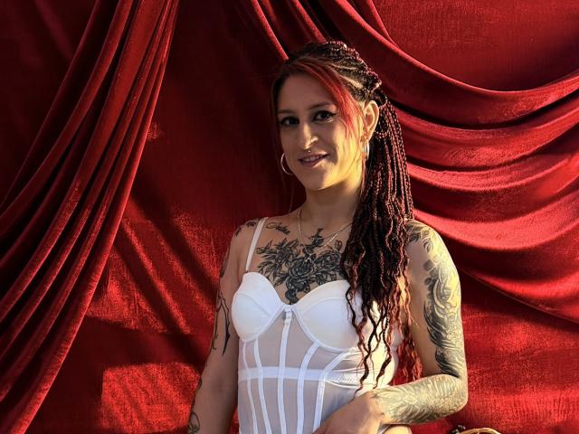 SofiFw with long red and black braided hair, white strapless lingerie, and tattoos, smiling at the camera. Red velvet backdrop. Boobs, tits, small tattoo on her neck.