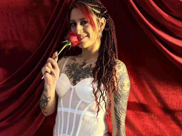 SofiFw with red rose, holding it to her mouth, has braided red and black hair, white sheer dress, tattoos on chest and arm, smiling, red velvet background.