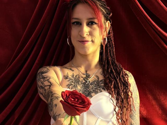 SofiFw with red braids, tattoos, nose ring, hoop earrings, holding red rose, white dress, smiling, red velvet background, light skin, medium breasts, and a necklace with a cross.