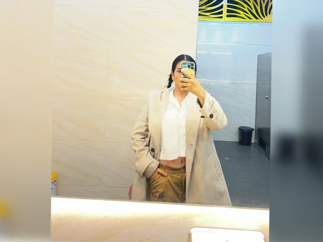 Velka with tan pants, white shirt, and beige coat, holding phone, mirror selfie, showing hand in coat pocket, black hair in braid, visible belly button.