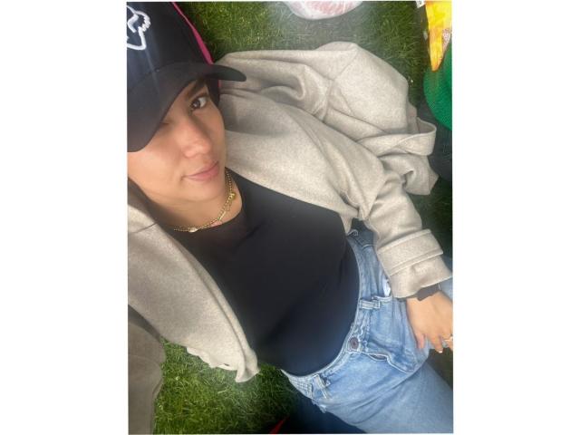 Velka with brown eyes, black cap, gold necklace, black top, grey jacket, and blue jeans, lying on green grass, looking up with a slight smile, showing her ass.