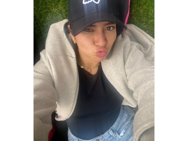 Velka with pouty lips, wearing a black cap, grey hoodie, black shirt, and blue jeans, has a gold necklace and is looking up at the camera.