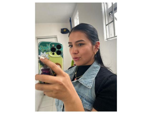 Velka with black hair, wearing a denim vest over a black shirt, taking a selfie with a green phone case in a white-walled room. She's pouting slightly.