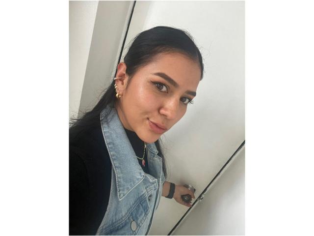 Velka with dark hair, wearing a denim vest, black shirt, and gold earrings, has a slight smirk, brown eyes, and a small nose, with a subtle smile.