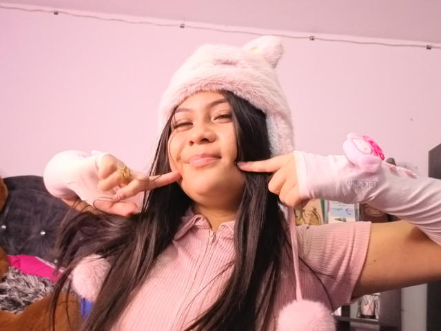 BadKristty with long black hair, wearing a pink fuzzy cat hat, pink ribbed shirt, and pink gloves, sticking her fingers near her cheek in a playful, confident pose.