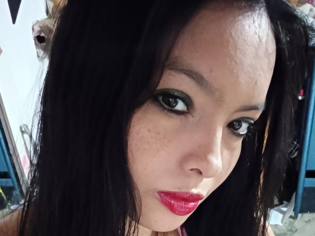 MelanieHot69's face with dark, wet-looking hair, green eyeshadow, and shiny pink lipstick. Her skin has freckles and a slightly sweaty forehead. She has a cute, sexy look.