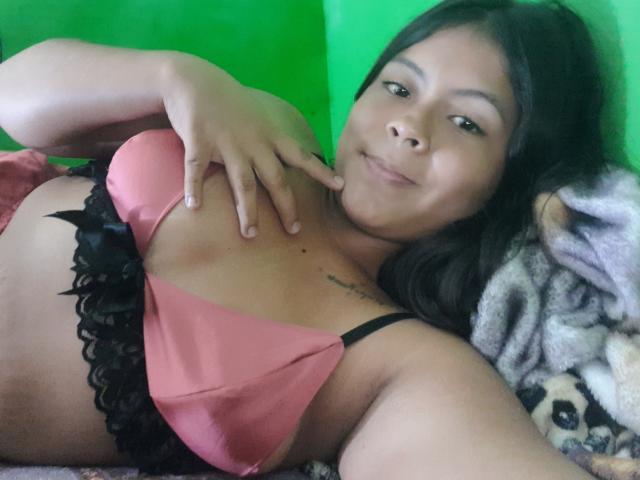 JulissaCortez lies on a bed, wearing a pink bra with black lace, her hand on her tit, smiling at the camera. Her dark hair and green background are visible.
