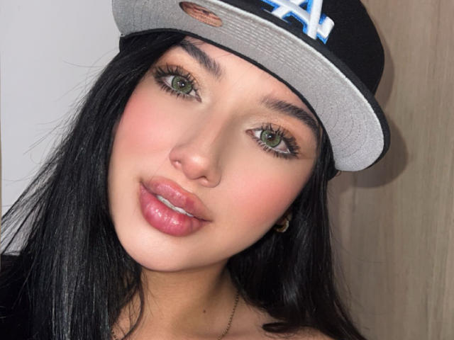 LillyTaylor with black hair, green eyes, pink glossy lips, wearing a black and grey cap, looking at the camera, has a cute and sexy face.