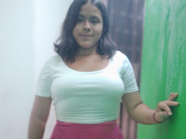 JulissaCortez with medium boobs, white t-shirt, pink high-waisted pants, dark brown wavy hair, red bracelet, green wall, slight smile, standing, right arm on wall.