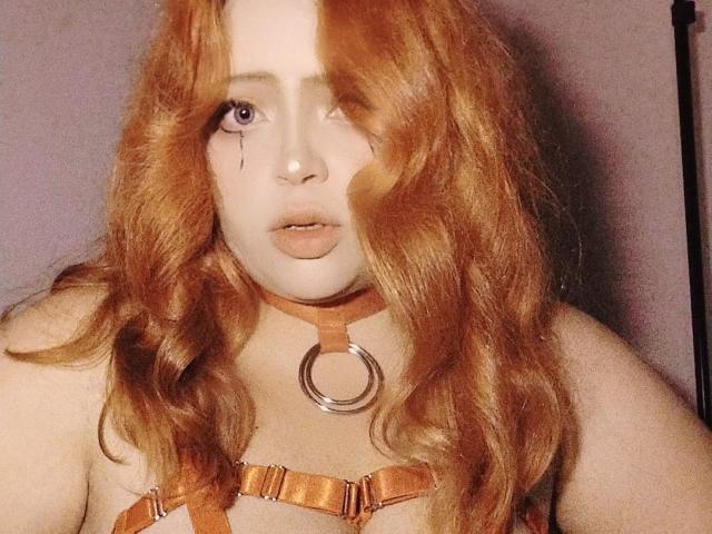 ChubbyLeahPearl with red wavy hair, pale skin, and a cute face, wearing an orange leather choker with a ring and matching harness, showing her boobs.
