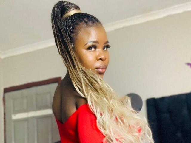 BustyAssHot with long blonde and black braids in a high pony, wearing a red off-shoulder top, looking to the side with a serious expression.