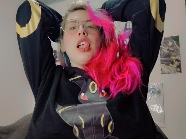NatashaLau with pink hair, glasses, tongue out, wearing black hoodie with Umbreon, arms raised, looking at camera, slightly messy, in a casual indoor setting.