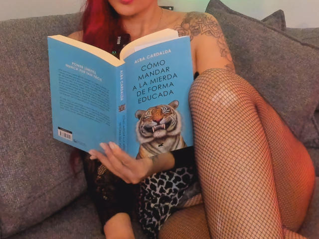 Laurent Colin mit roten Haaren, Tätowierungen, Netzstrümpfen, einem Leopardenmuster-Kleid, liest ein Buch mit einem Tiger auf dem Cover, auf einem grauen Sofa. Brüste sind teilweise sichtbar, Buch in der Hand, entspannt, sexy..