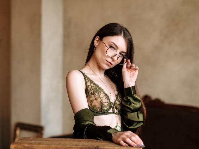 MelanieFoster with long dark hair, green lace bra, and glasses, adjusting them with her right hand, wearing an off-shoulder green top, standing by a wooden table.