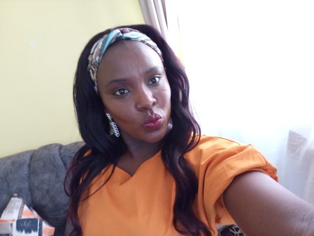 CammieBobby with dark brown skin, long wavy hair, wearing an orange shirt, patterned headband, and dangling earrings, pouting her red lips, standing in front of a window.