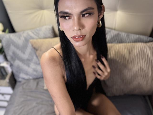 CassandraWise has long black hair, light brown skin, and is wearing a black, low-cut top. She has full lips with pink lipstick, dark eyebrows, and brown eyes. She's looking up with a slightly open mouth. She has small, white ear gauges and black nail polish. She's sitting on a bed with gray, patterned pillows and beige, tufted headboard. The focus is on her upper body and face. Her right hand is touching her hair. The photo has a shallow depth of field, making the background blurry.