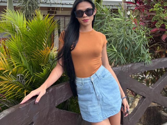 CassandraWise with long black hair, brown t-shirt, blue denim skirt, black sunglasses, and red lipstick, leaning on a wooden fence with green and red plants behind her.