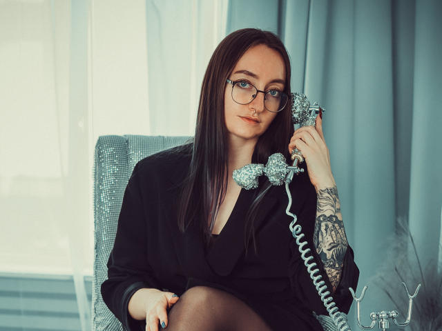 Shiroheby con gafas, vestido negro y tatuajes, sosteniendo un teléfono de plata antiguo, sentada en una silla gris, mirando a la cámara, con cortinas azules de fondo..