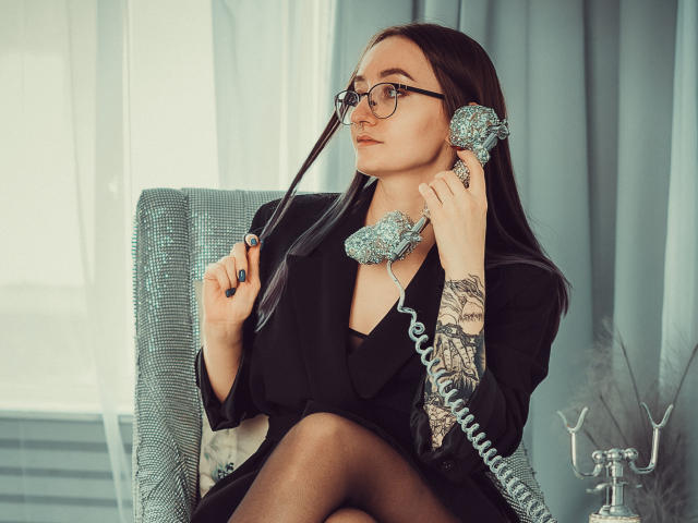 Shiroheby, con gafas, un blazer negro y un teléfono azul, sosteniendo el auricular en su oído, tiene tatuajes en su brazo izquierdo y está sentada en una silla color turquesa..