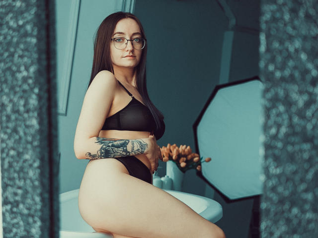 Shiroheby, con cabello castaño oscuro, gafas, un sostén y ropa interior negros, mostrando un tatuaje en su brazo derecho, sentada en un taburete blanco, mirando a la cámara..