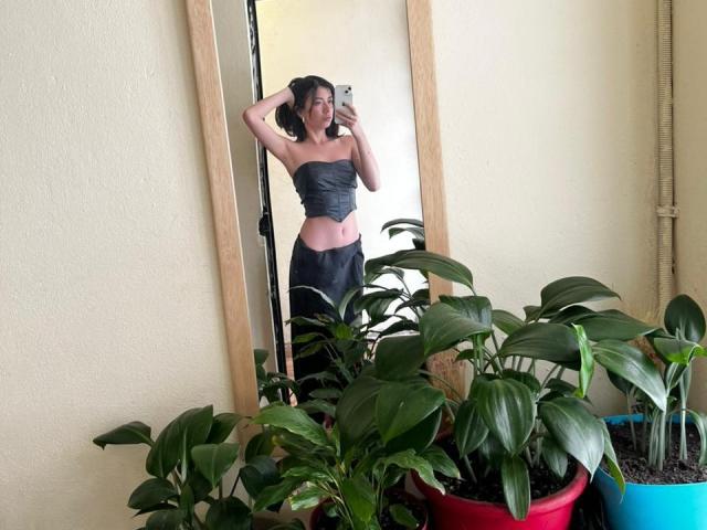 AnyuWu, com cabelo escuro, vestindo uma blusa cinza sem alças, mostrando a cintura fina, está em frente a um espelho, cercada por plantas em vasos verdes..