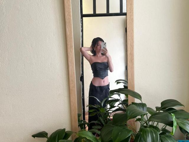 AnyuWu macht ein Selfie vor einem Spiegel, trägt ein schwarzes Top ohne Träger und einen schwarzen Rock, eine Hand liegt auf ihrem Kopf, und sie zeigt ihren flachen Bauch und ihre kleinen Brüste, umgeben von grünen Pflanzen..