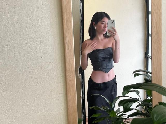 AnyuWu trägt ein schwarzes, trägerloses Korsett, das ihre Brüste zeigt, und schwarze Hosen. Sie macht ein Selfie vor einem Spiegel, wobei ihre Hand auf ihrer Brust liegt. Vor ihr steht eine grüne Pflanze..