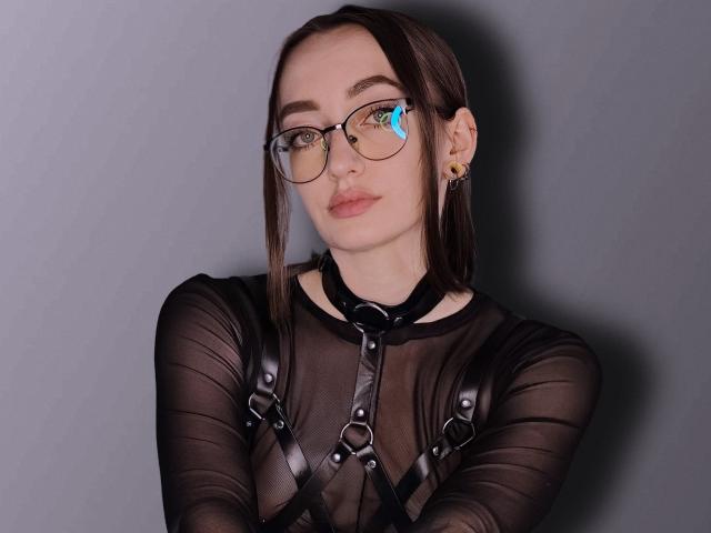 Shiroheby, con gafas, lleva puesto un top negro transparente y un arnés de cuero, tiene una expresión seria, cabello castaño y un arete dorado, mirando directamente a la cámara..