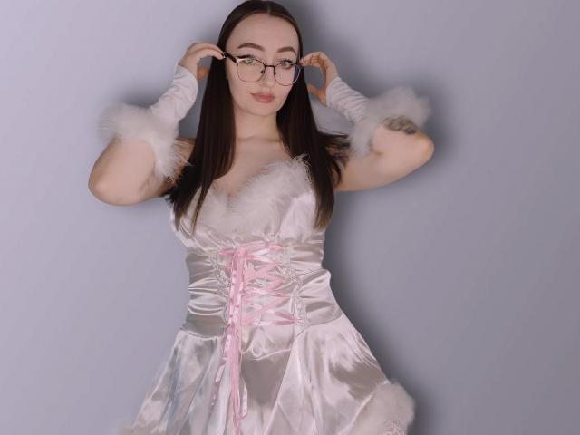 Shiroheby, con cabello largo y castaño, gafas, puños de piel blanca, un vestido de satén blanco con cordones rosas y guantes blancos, ajustándose las gafas con las manos. Tiene tatuajes en su brazo derecho..