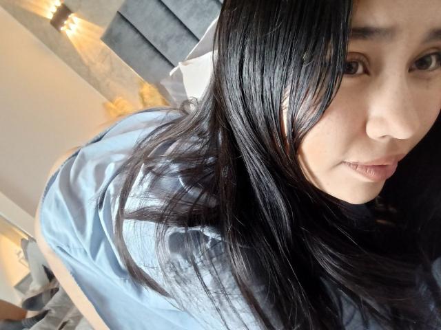 Karollteyes with long black hair, wearing a light blue shirt, showing her ass, looking at the camera with a slight smile, in a bedroom.