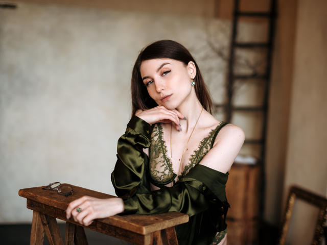 MelanieFoster with pale skin, dark brown hair, green lace bra, off-shoulder green satin robe, hand on wooden table, ladder in background, looking at camera, earrings.