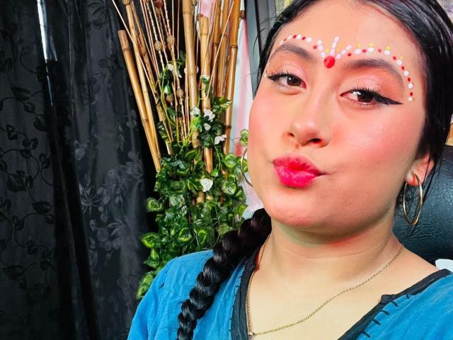 KalpanaAisha has pink lipstick, a red dot on her forehead, braid, blue top, hoop earrings, and colorful eye makeup with dots. Her ass is not visible.
