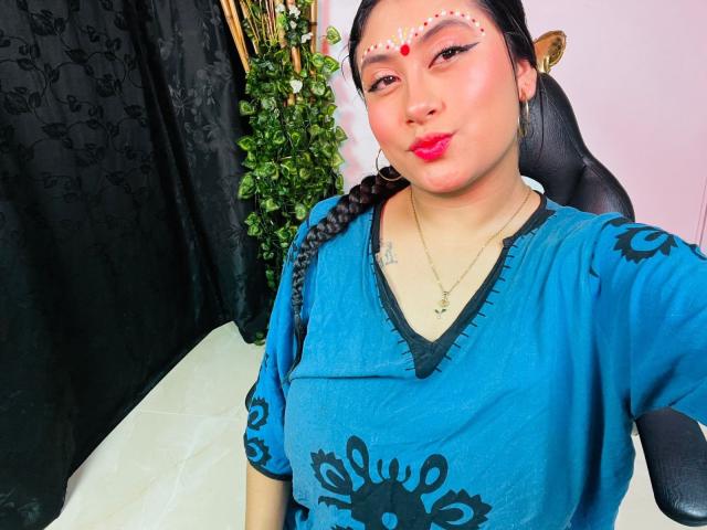 KalpanaAisha with braided hair, blue patterned top, red dot makeup, hoop earrings, pink lipstick, and necklace, pouting, black curtain, green plant, and brown statue in background.