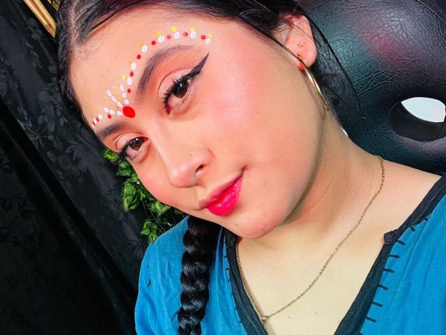 KalpanaAisha has colorful face paint with red, white, and yellow dots above her eyebrows, and a braid in her dark hair, wearing a blue top and gold earrings.