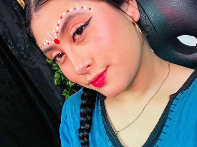 KalpanaAisha with colorful face paint, braid, and blue top, showing off her red lips and gold hoop earrings, looking cute and confident. Her tits are not visible.