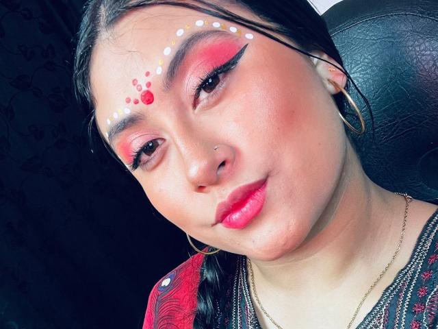 KalpanaAisha's face with bright pink and red eyeshadow, white and red dots on her forehead, black eyeliner, pink lips, nose piercing, and gold hoop earrings, smiling slightly.