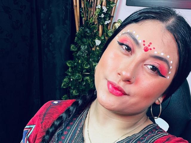 KalpanaAisha has red eye makeup, white dots on her forehead, and pink lips. She's wearing a red patterned shirt, hoop earrings, and a nose piercing. Black hair in a braid.