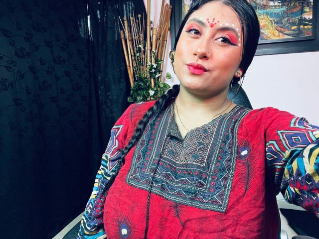 KalpanaAisha with medium tits, red dress, colorful embroidery, black braids, red makeup, white facial dots, black curtain, decorative wall, and wooden sticks in the background.